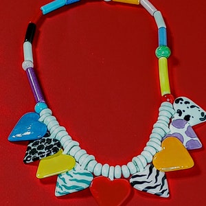 May include: A colorful beaded necklace with a variety of heart-shaped charms. The charms are decorated with different patterns, including zebra stripes, polka dots, and animal print. The necklace is made with white, yellow, blue, green, red, and purple beads.