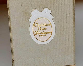 May include: A small, rectangular box with a light gray cardboard exterior. The box has a gold trim around the top and bottom edges. A white oval sticker with a gold trim and a gold bow is centered on the front of the box. The sticker reads "Christian Dior Diorissima Paris".