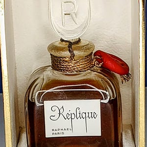 May include: A vintage glass bottle of Replique perfume by Raphael Paris. The bottle has a clear glass stopper with a gold-colored R on it. The bottle is filled with a dark brown liquid and has a red wax seal on the top.