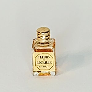 May include: A small, rectangular perfume bottle with a gold-colored cap and label. The label reads "FLEURS DE ROCAILLE CANON" in a gold octagonal frame. The bottle contains a light amber liquid.