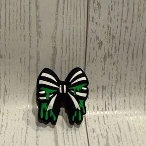 May include: A black and white bow-shaped charm featuring a dripping green slime effect. The design consists of a central knot with two loops and two tails, all rendered in a high-contrast black and white color scheme with bright green accents.