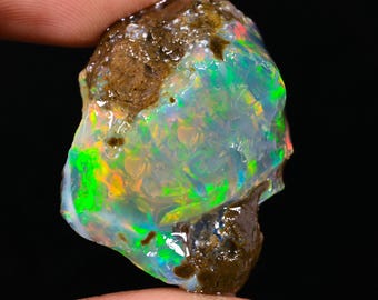 May include: A rough opal gemstone with a vibrant play of color, including green, blue, and orange hues. The opal is held between fingers, showcasing its natural, unpolished texture and earthy brown matrix. The gemstone is set against a dark background.