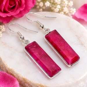 May include: A pair of drop earrings featuring rectangular, deep pink, faceted stones set in silver-toned metal frames. Each earring is attached to a simple silver-toned hook wire, showcasing a polished finish and a vibrant, saturated color.