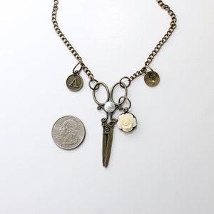 May include: A gold necklace with a pair of antique-style scissors, a pearl, a white rose, and two charms: one with the letter 'A' and one with a button design.