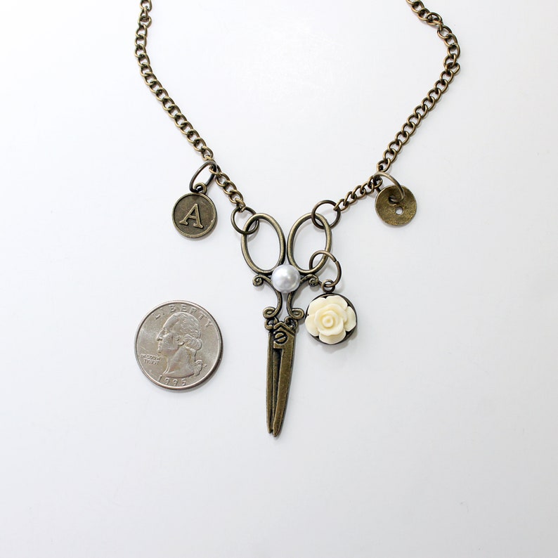 May include: A gold necklace with a pair of antique-style scissors, a pearl, a white rose, and two charms: one with the letter 'A' and one with a button design.