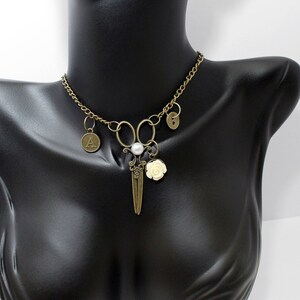 May include: A bronze necklace with a pair of scissors charm, a pearl, a white rose charm, and two letter charms. The necklace is on a black mannequin.