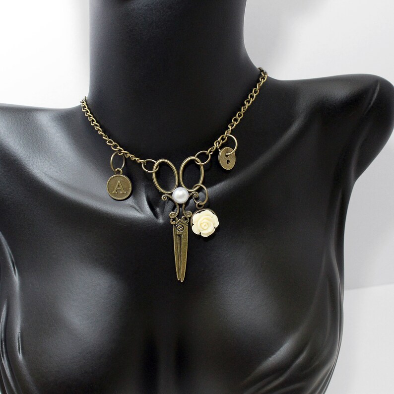 May include: A bronze necklace with a pair of scissors charm, a pearl, a white rose charm, and two letter charms. The necklace is on a black mannequin.