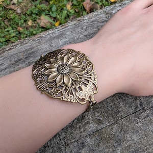 Vintage brass sunflower bracelet with leaf detail displayed on wrist over a log outdoors.