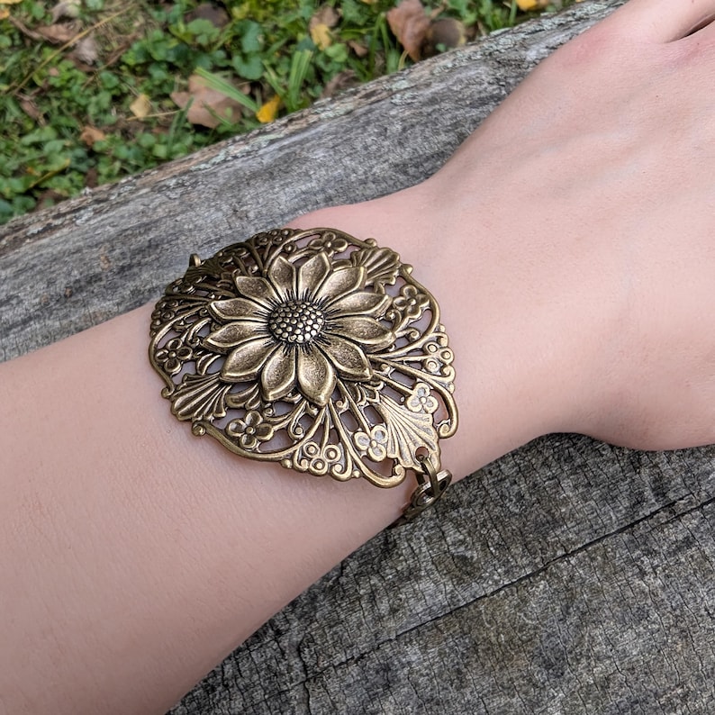 Vintage brass sunflower bracelet with leaf detail displayed on wrist over a log outdoors.