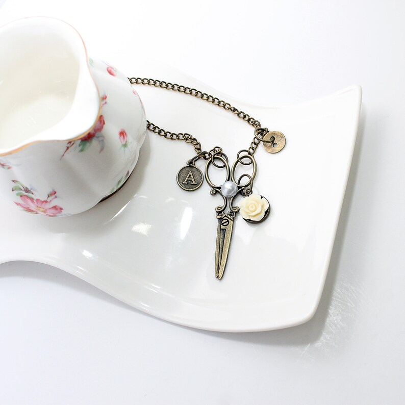 May include: A gold-toned necklace with a charm of a pair of scissors, a pearl, a small white flower, and a letter 'A'.