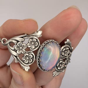 Può includere: Una collana con pendente color argento con una gemma di opale ovale dai colori iridescenti. Il pendente è incorniciato da un motivo a corda intrecciata e da dettagli floreali in filigrana. La collana è tenuta da una mano.