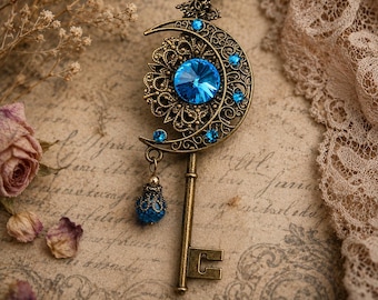 May include: A vintage-style key pendant necklace. The pendant features a crescent moon design with intricate filigree and a large, round, blue gemstone at the center. Smaller blue gemstones accent the moon. The key is a bronze color, and the necklace has a matching chain.