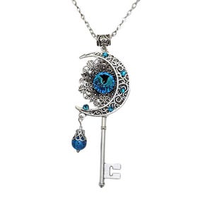 Silver-toned pendant necklace featuring a crescent moon design with intricate filigree and blue gemstone accents. The pendant includes a key shape and a dangling blue bead. The necklace is suspended from a delicate chain.