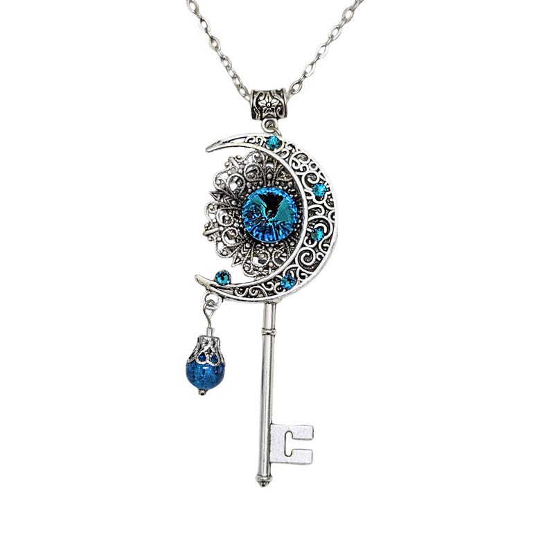 Silver-toned pendant necklace featuring a crescent moon design with intricate filigree and blue gemstone accents. The pendant includes a key shape and a dangling blue bead. The necklace is suspended from a delicate chain.
