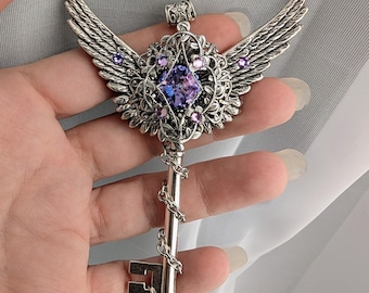 May include: A silver-colored key pendant with a wing design. The key features a large, purple gemstone at its center, surrounded by smaller purple gems and intricate floral detailing. A chain wraps around the key's stem.