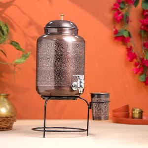 May include: A copper-colored water dispenser with a decorative floral pattern, a spigot, and a matching cup. The dispenser sits on a black metal stand. The background is a warm orange.