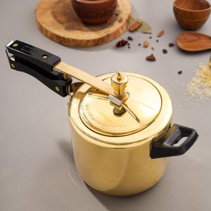 May include: A gold-colored pressure cooker with a black handle and lid. The cooker has a shiny finish and a pressure release valve on top. The handle is made of black plastic and is attached to the lid with a gold-colored metal arm.