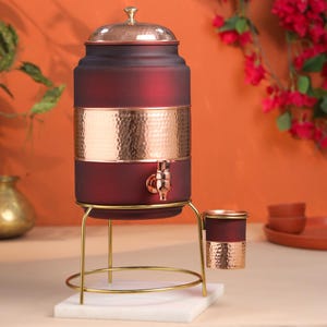 Pode incluir: Um dispensador de bebidas bordô e cobre com torneira, tampa e detalhes em metal martelado. Ele fica em um suporte de metal dourado com um porta-copos correspondente. O dispensador está em uma base de mármore branco.