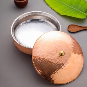 Può includere: Una ciotola da portata color rame con una texture martellata e un coperchio abbinato. Il coperchio è parzialmente aperto, rivelando l'interno argentato della ciotola. Un piccolo pomello dorato adorna il coperchio. Sono visibili anche un cucchiaio di legno e una foglia verde.