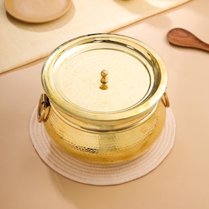 May include: A gold-coloured, hammered-metal cooking pot with a lid and two handles. The pot sits on a round, off-white woven coaster. A wooden spoon and a plate are visible in the background.