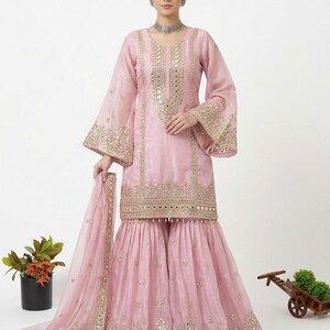 Puede incluir: Un salwar kameez rosa claro con bordados dorados y detalles de espejo. El conjunto incluye un dupatta a juego y pantalones de pernera ancha. Las mangas tienen forma de campana. Este conjunto es adecuado para ocasiones especiales.