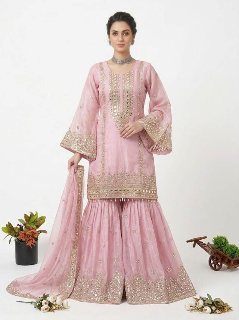 Puede incluir: Un salwar kameez rosa claro con bordados dorados y detalles de espejo. El conjunto incluye un dupatta a juego y pantalones de pernera ancha. Las mangas tienen forma de campana. Este conjunto es adecuado para ocasiones especiales.