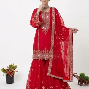 Puede incluir: Un vibrante conjunto rojo con una t&uacute;nica de manga larga y pantalones de pernera ancha, adornado con intrincados bordados dorados y espejos. Una bufanda transparente a juego completa el atuendo. Prenda tradicional india para ocasiones festivas.