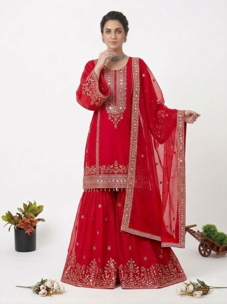 Puede incluir: Un vibrante conjunto rojo con una t&uacute;nica de manga larga y pantalones de pernera ancha, adornado con intrincados bordados dorados y espejos. Una bufanda transparente a juego completa el atuendo. Prenda tradicional india para ocasiones festivas.