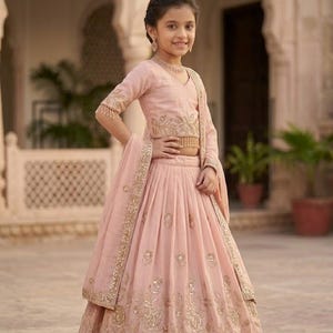 Peut inclure: Un ensemble lehenga choli rose p&acirc;le compos&eacute; d'un haut court &agrave; manches trois-quarts et d'une jupe pliss&eacute;e. Les deux pi&egrave;ces sont orn&eacute;es de broderies dor&eacute;es complexes et de petits miroirs r&eacute;fl&eacute;chissants. Un dupatta assorti est drap&eacute; sur l'&eacute;paule.