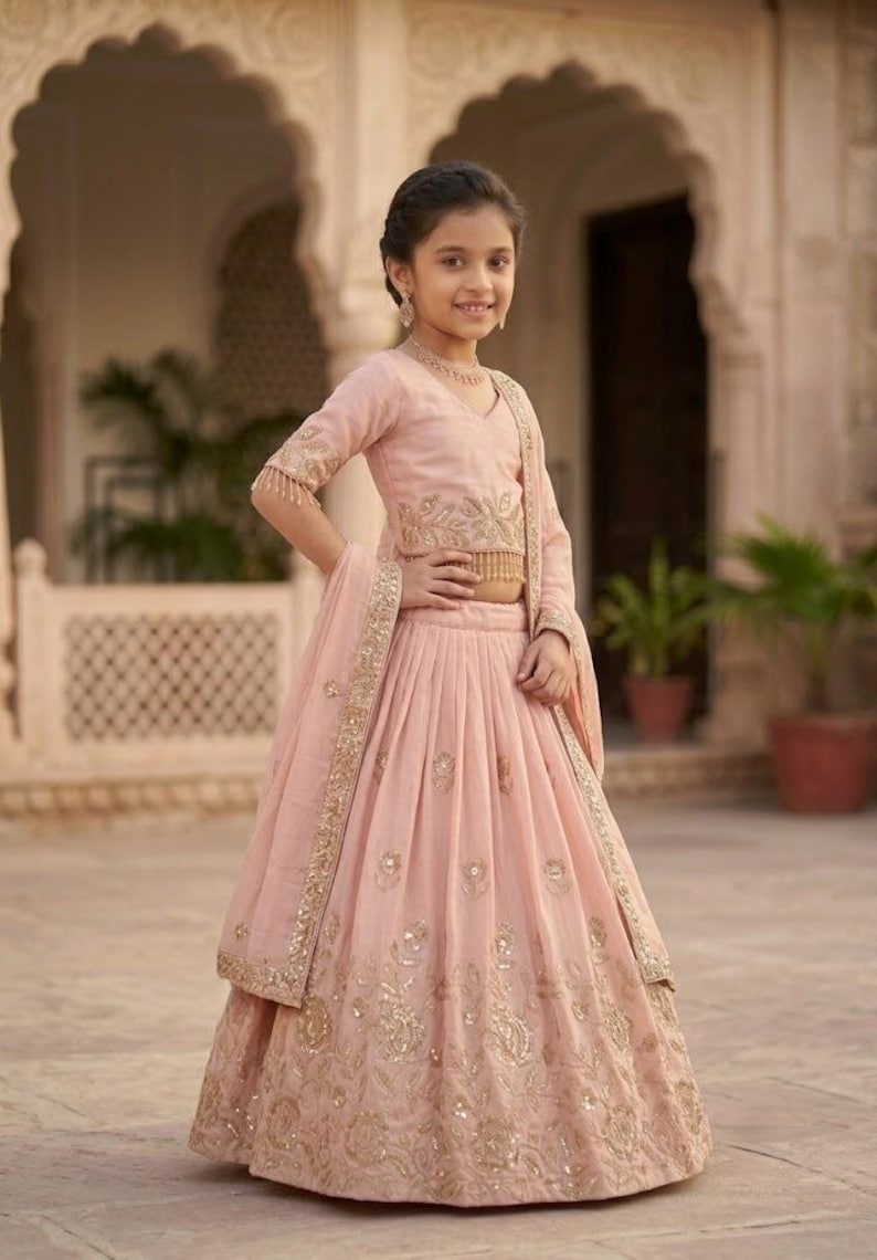 Peut inclure: Un ensemble lehenga choli rose p&acirc;le compos&eacute; d'un haut court &agrave; manches trois-quarts et d'une jupe pliss&eacute;e. Les deux pi&egrave;ces sont orn&eacute;es de broderies dor&eacute;es complexes et de petits miroirs r&eacute;fl&eacute;chissants. Un dupatta assorti est drap&eacute; sur l'&eacute;paule.