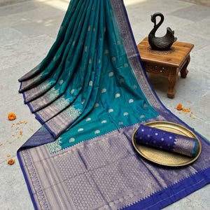 Könnte beinhalten: Ein türkis- und marineblauer Sari mit aufwendigen silberfarbenen Mustern. Der Sari ist drapiert und zeigt seinen detaillierten Rand und sein Design. Ein aufgerollter Teil des Saris liegt auf einem goldfarbenen Tablett.