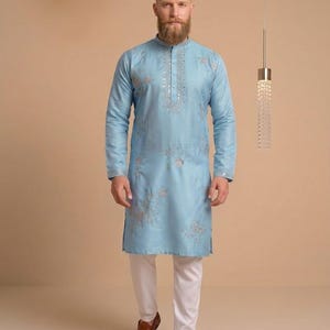 Puede incluir: Un kurta azul claro con bordados intrincados y cuello mandarín, combinado con pantalones blancos. La parte superior de manga larga presenta adornos plateados detallados. El atuendo se completa con mocasines de cuero marrón.