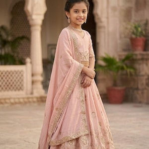Peut inclure: Un ensemble lehenga choli rose clair compos&eacute; d'un haut sans manches et d'une jupe longue, tous deux orn&eacute;s de broderies dor&eacute;es et de sequins. Un dupatta assorti en voile avec une bordure brod&eacute;e dor&eacute;e est drap&eacute; sur l'&eacute;paule.