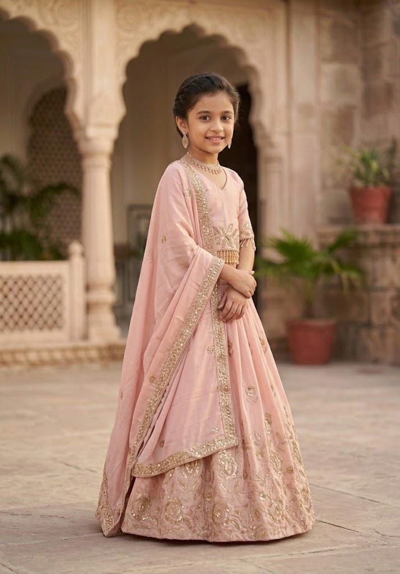 Peut inclure: Un ensemble lehenga choli rose clair compos&eacute; d'un haut sans manches et d'une jupe longue, tous deux orn&eacute;s de broderies dor&eacute;es et de sequins. Un dupatta assorti en voile avec une bordure brod&eacute;e dor&eacute;e est drap&eacute; sur l'&eacute;paule.
