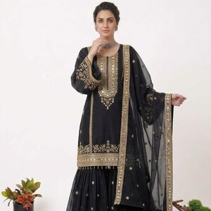 Puede incluir: Conjunto salwar kameez negro bordado. El atuendo presenta intrincados bordados dorados en el escote, las mangas y el dobladillo. Un dupatta transparente a juego con ribete dorado completa el look. El atuendo lo lleva una persona.
