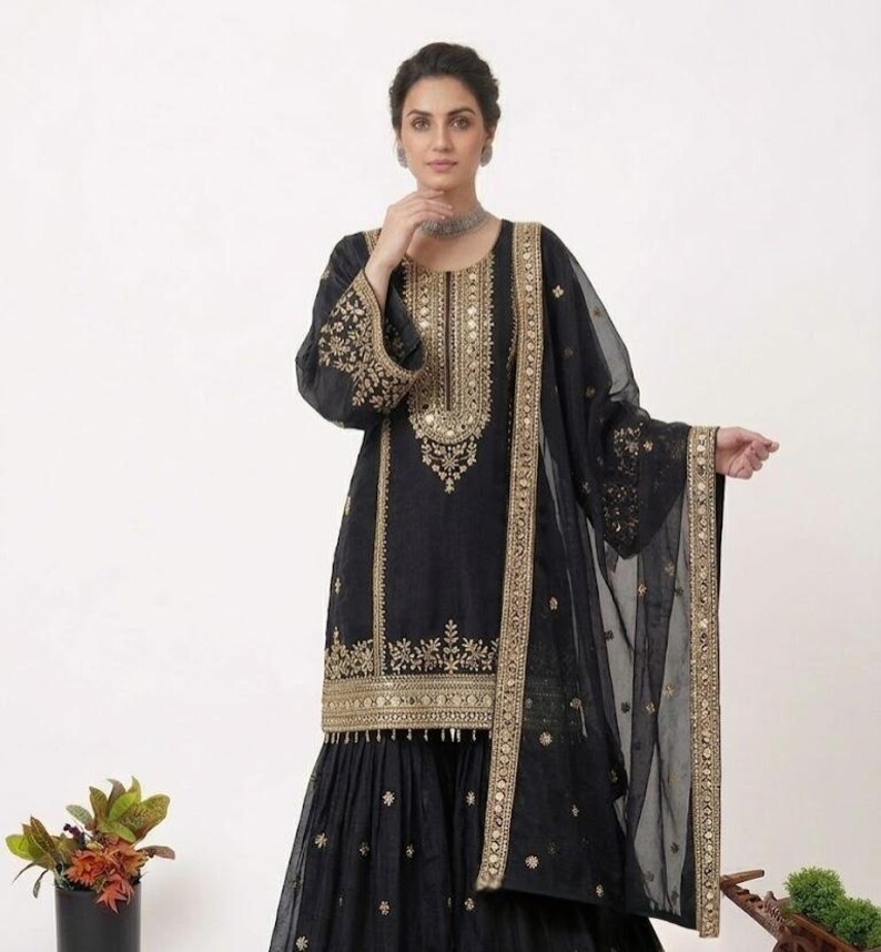 Puede incluir: Conjunto salwar kameez negro bordado. El atuendo presenta intrincados bordados dorados en el escote, las mangas y el dobladillo. Un dupatta transparente a juego con ribete dorado completa el look. El atuendo lo lleva una persona.