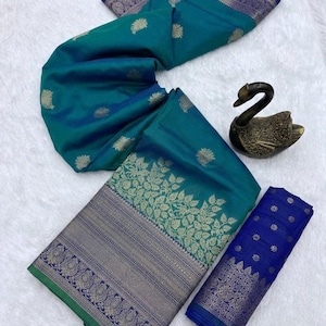 May include: Teal saree with gold floral detailing and a navy blue border. The fabric is displayed on a white surface, highlighting the intricate design. A separate piece of matching fabric is also visible, showcasing the rich texture.