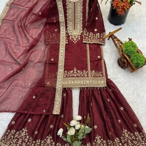 Puede incluir: Conjunto salwar kameez burdeos con bordados dorados. El conjunto incluye una t&uacute;nica de manga larga, pantalones de pernera ancha y una bufanda transparente. La t&uacute;nica y los pantalones presentan intrincados dise&ntilde;os florales y geom&eacute;tricos.