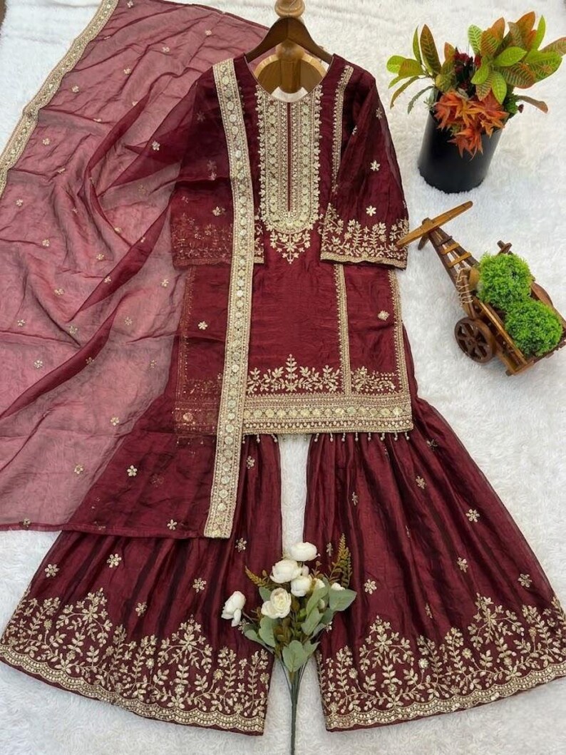 Puede incluir: Conjunto salwar kameez burdeos con bordados dorados. El conjunto incluye una t&uacute;nica de manga larga, pantalones de pernera ancha y una bufanda transparente. La t&uacute;nica y los pantalones presentan intrincados dise&ntilde;os florales y geom&eacute;tricos.