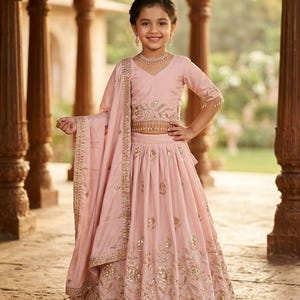 Peut inclure: Un ensemble lehenga choli rose clair compos&eacute; d'un haut court &agrave; manches courtes et d'une jupe longue assortie. Les deux pi&egrave;ces sont orn&eacute;es de broderies dor&eacute;es complexes et de motifs floraux &agrave; sequins, avec un dupatta drap&eacute;.