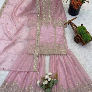 Puede incluir: Conjunto salwar kameez rosa claro bordado en dorado. El conjunto incluye una t&uacute;nica de manga larga, pantalones de pernera ancha y una bufanda a juego. La t&uacute;nica y los pantalones est&aacute;n adornados con bordados dorados. El conjunto se muestra sobre una superficie blanca.