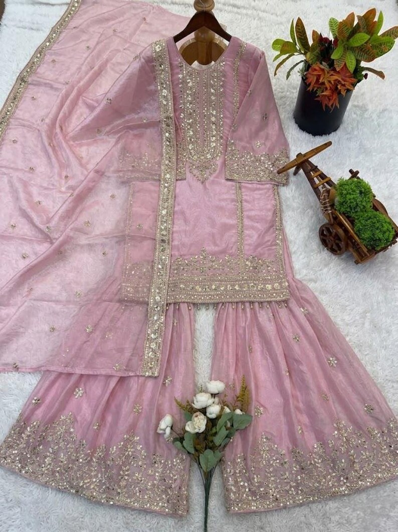 Puede incluir: Conjunto salwar kameez rosa claro bordado en dorado. El conjunto incluye una t&uacute;nica de manga larga, pantalones de pernera ancha y una bufanda a juego. La t&uacute;nica y los pantalones est&aacute;n adornados con bordados dorados. El conjunto se muestra sobre una superficie blanca.