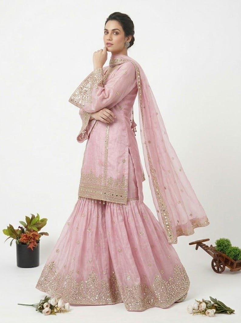 Puede incluir: Un traje sharara rosa claro con adornos dorados. El conjunto incluye una parte superior de manga larga, una falda acampanada y una dupatta a juego. Las mangas y los dobladillos de la falda est&aacute;n detallados con bordados dorados y espejos.