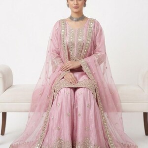 Puede incluir: Conjunto salwar kameez rosa claro adornado. El atuendo incluye una t&uacute;nica de manga larga, pantalones de pernera ancha y un dupatta transparente. La t&uacute;nica y los pantalones presentan intrincados bordados dorados y espejos. La modelo lleva un collar y pendientes plateados.
