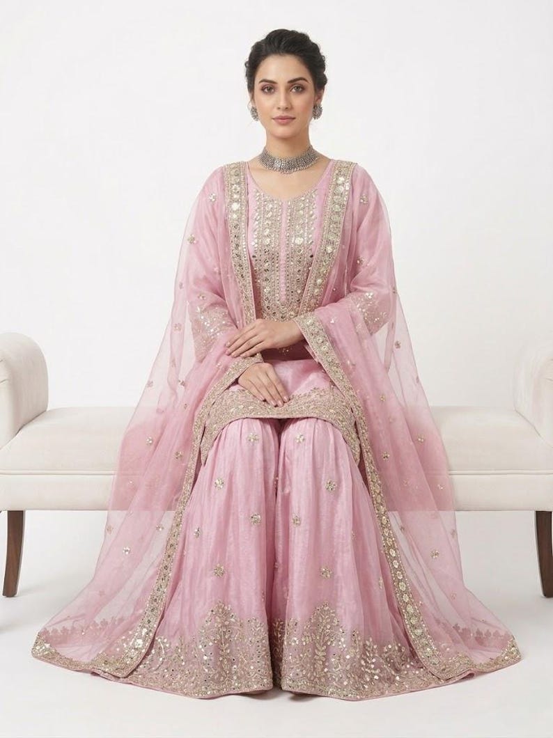 Puede incluir: Conjunto salwar kameez rosa claro adornado. El atuendo incluye una t&uacute;nica de manga larga, pantalones de pernera ancha y un dupatta transparente. La t&uacute;nica y los pantalones presentan intrincados bordados dorados y espejos. La modelo lleva un collar y pendientes plateados.