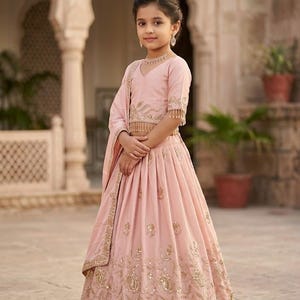 Peut inclure: Un ensemble lehenga choli rose clair compos&eacute; d'un chemisier &agrave; manches courtes avec broderies dor&eacute;es et franges, assorti d'une jupe pliss&eacute;e orn&eacute;e de motifs floraux dor&eacute;s complexes et d'un dupatta coordonn&eacute; drap&eacute; sur l'&eacute;paule.