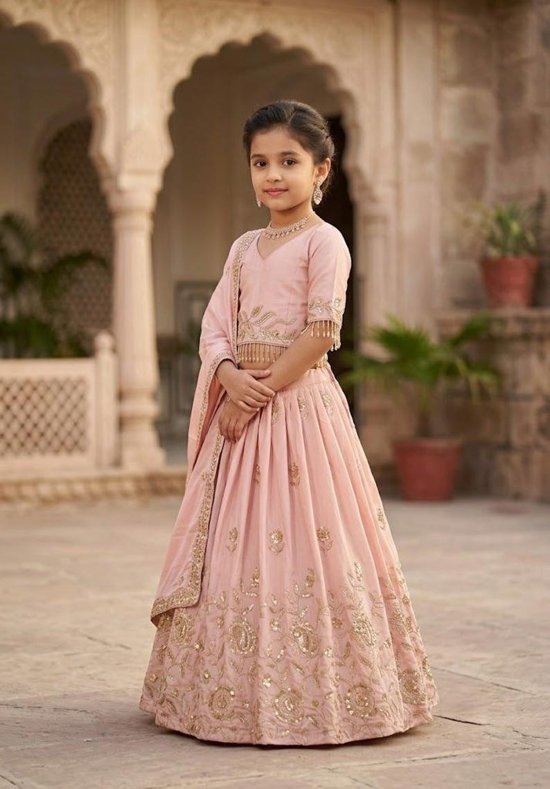 Peut inclure: Un ensemble lehenga choli rose clair compos&eacute; d'un chemisier &agrave; manches courtes avec broderies dor&eacute;es et franges, assorti d'une jupe pliss&eacute;e orn&eacute;e de motifs floraux dor&eacute;s complexes et d'un dupatta coordonn&eacute; drap&eacute; sur l'&eacute;paule.