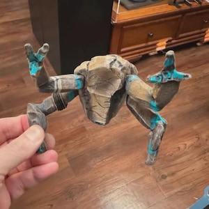 May include: A small, articulated figurine resembling a rock-like creature with a textured, tan-colored central body and four limbs. The limbs feature a weathered gray finish with bright turquoise accents at the joints and claw-like tips.