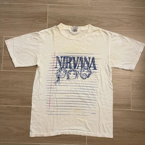 Può includere: Una t-shirt bianca a maniche corte con una stampa grafica blu della parola NIRVANA sopra tre volti disegnati a mano. Il design è posto su uno sfondo di linee orizzontali blu che ricordano la carta da quaderno con una linea di margine rossa.