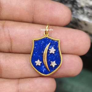 May include: A gold-colored pendant with a shield shape, featuring a deep blue background with a gold crescent moon and three star-shaped accents. The pendant has a textured border and a small loop for a chain.